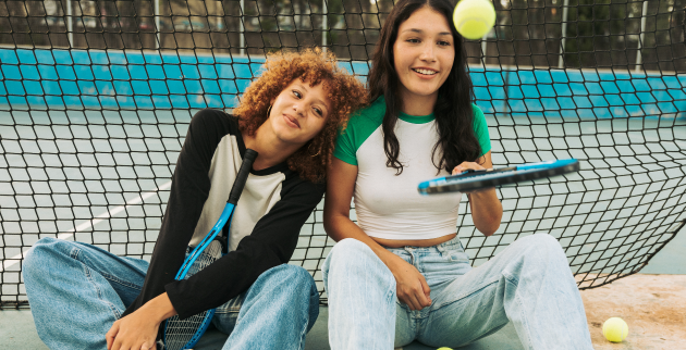Two friends sat down on a tennis court. One is bouncing a tennis ball on a tennis racket.